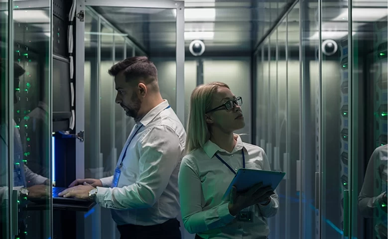 Two people in white button-ups holding tablets and walking around a server room.