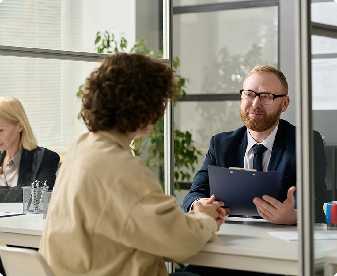 A man in a suit holds a clipboard and speaks to a woman in a beige jacket.